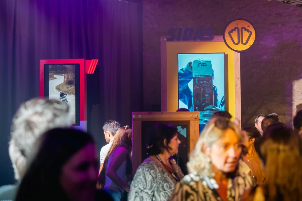Crowd of people at an indoor exhibit with colorful display screens and a yellow-framed panel on a textured wall.
