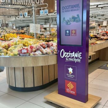 Purple Occitanie promotional display in a supermarket produce area, with fresh fruit in the foreground and branding reading 'Occitanie, tu nous régales'