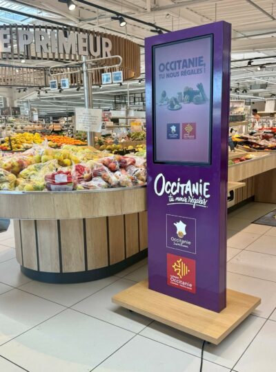 Purple Occitanie promotional display in a supermarket produce area, with fresh fruit in the foreground and branding reading 'Occitanie, tu nous régales'